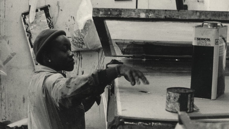 Black and white photograph of the artist Claudette Johnson, dressed in overalls, using screen-printing equipment at the Lenthall Road Workshop.