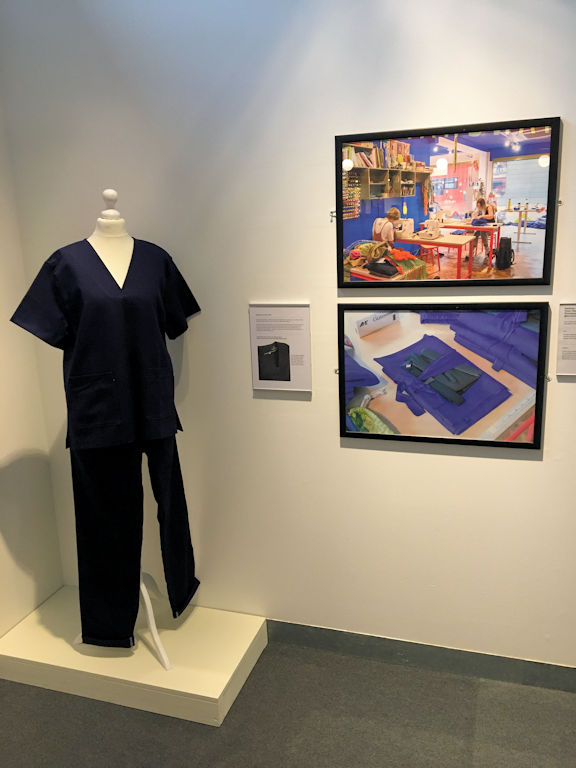 view of a museum exhibition. A mannequin wearing surgical scrubs is next to two framed images showing how the items were made.