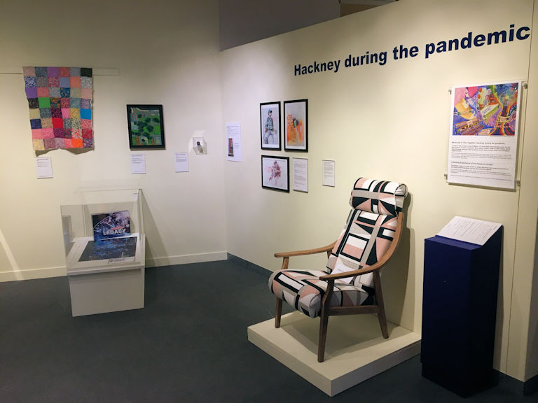view of a museum exhibition. Display cases, framed images and a chair are displayed nuder the heading 'Hackney During the Pandemic'