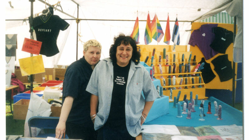 Colour photograph of two women (one is Gonul Zeki) posing in a market stall decorated with rainbow flags selling t-shirts with LGBTQI+ slogans