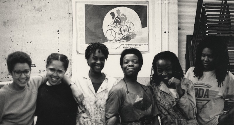 Black and white photograph of six women embracing one another and smiling at a fabric printing workshop
