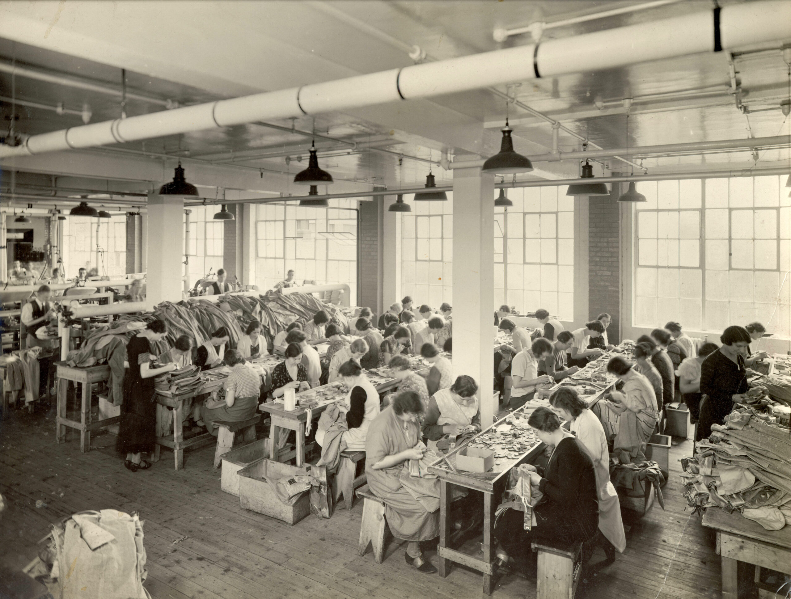 Sepia image of all-female workforce at the Simpsons Clothing Factory. The women are sitting down at long wooden benches, facing one another and working on garments.