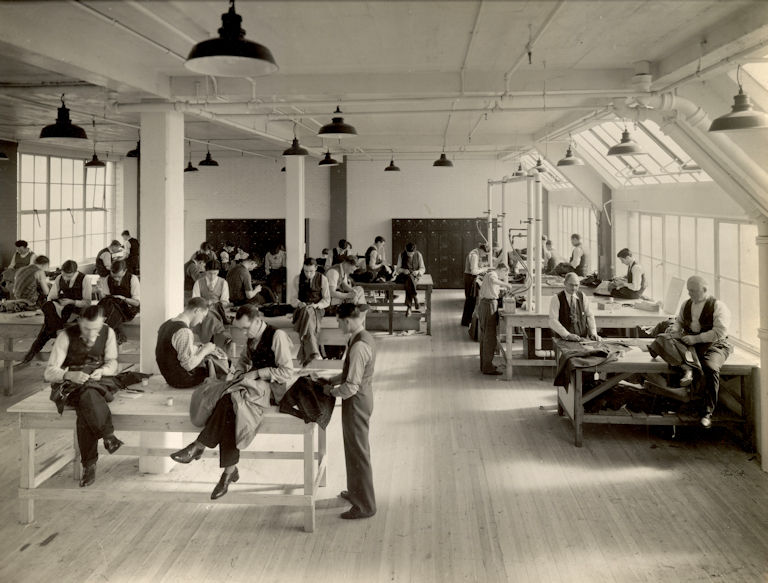 Sepia image of garment workers inside the Simpsons Clothing factory. There are lots of smartly dressed men who are working on garments by hand. Some are sitting on workbenches and others are standing.