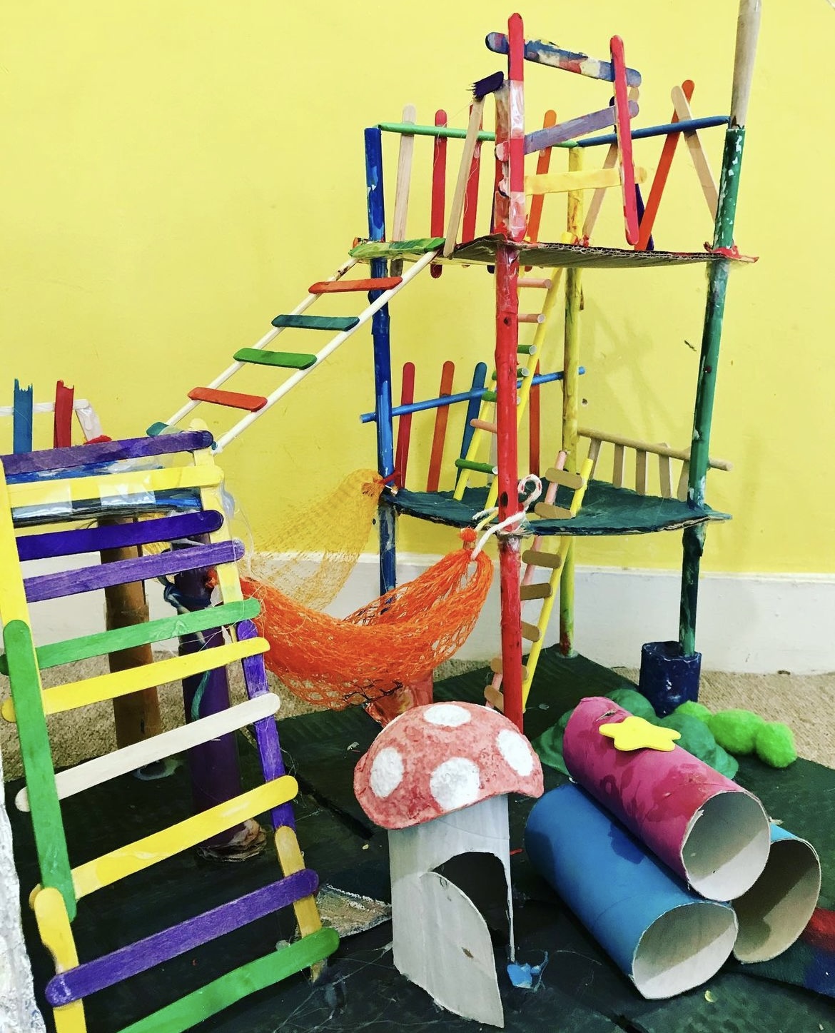 Colorful model of an adventure playground made out of junk materials e.g. ladders made of ice lolly sticks, hammock made of tangerine netting and mushroom made of toilet roll tube