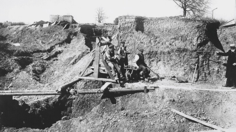 A black and white photograph of a deep excavation. In the trench pose 3 unknown workers, and apart from them Worthington George Smith pointing to a layer of stratigraphy in the soil with his cane.