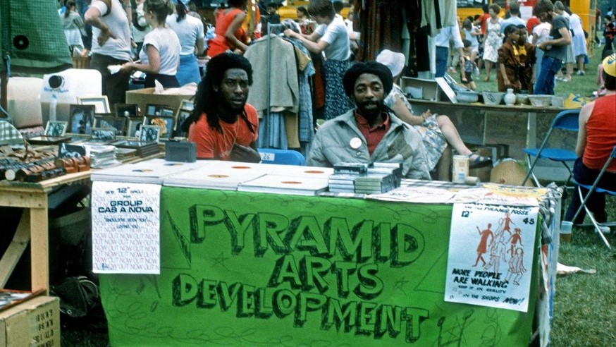 A colour photograph of a busy community event in a field, with a number of stalls in the foreground. The photo is focussed on one stall with a large green banner reading "Pyramid Arts Development" supervised by two men of African heritage. The stall is selling records and cassette tapes.