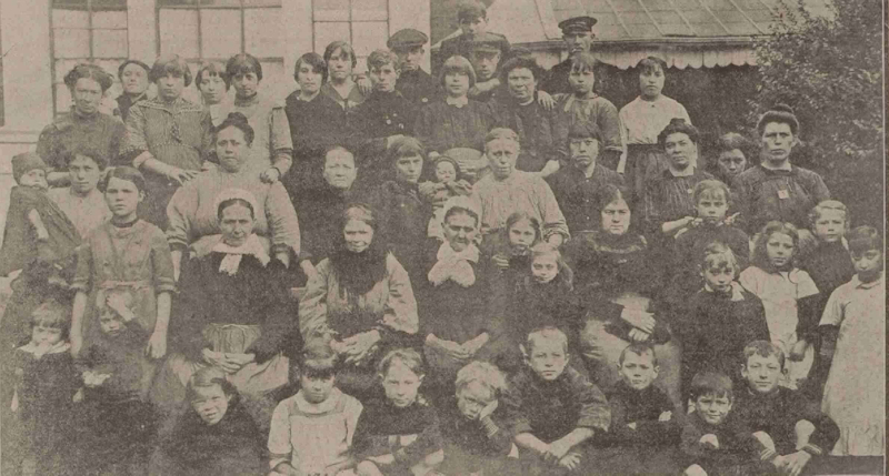 A sepia photo of around 50 women, children and men arranged for a group photo, wearing Edwardian clothing. Belgian Refugees in Hackney during the First World War. 