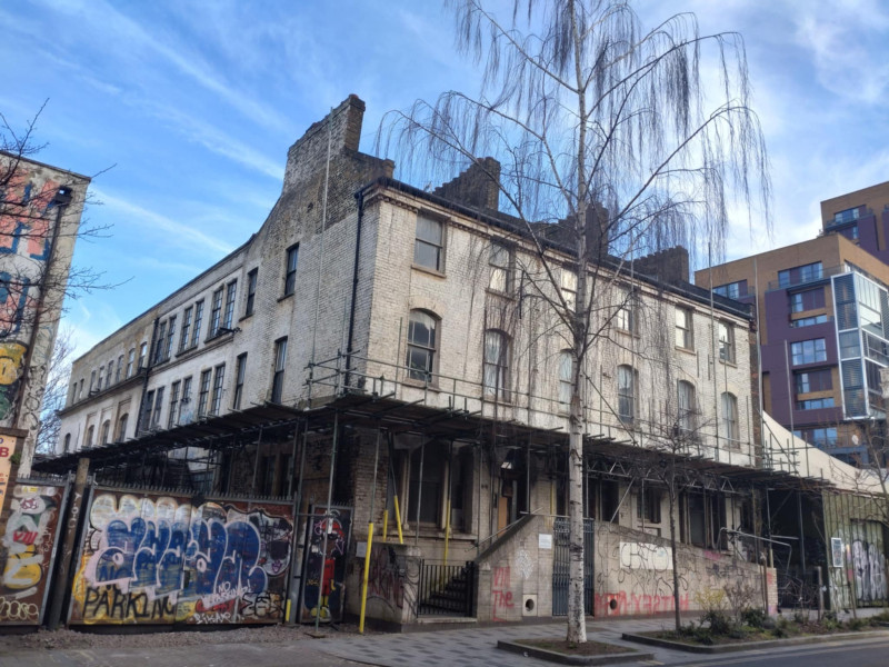 A photograph of 10-16 Ashwin Street. It shows a terrace of white 3 storey properties with a large industrial style warehouse space behind. The property has scaffolding and some graffiti.
