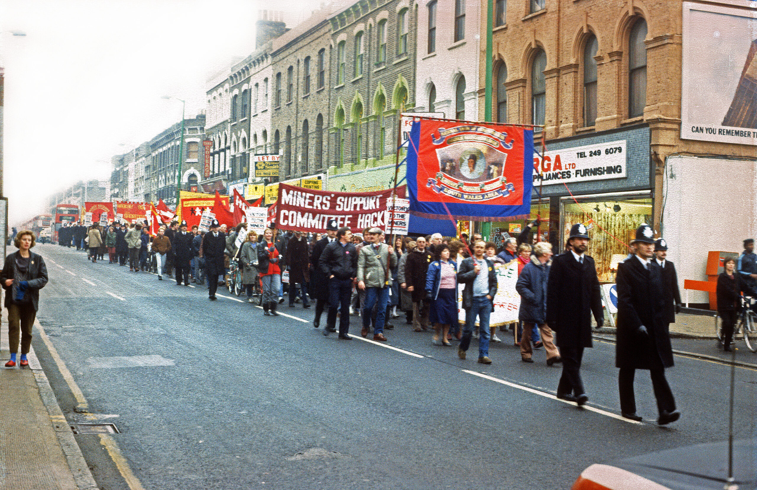 A colour photograph of a demonstration marching down Kingsland High Street, Hackney, in front of Centerprise Bookshop, with a police officer walking in front. Marchers hold banners for the National Union of Mineworkers South Wales Area, and the Miners Support Committee Hackney.