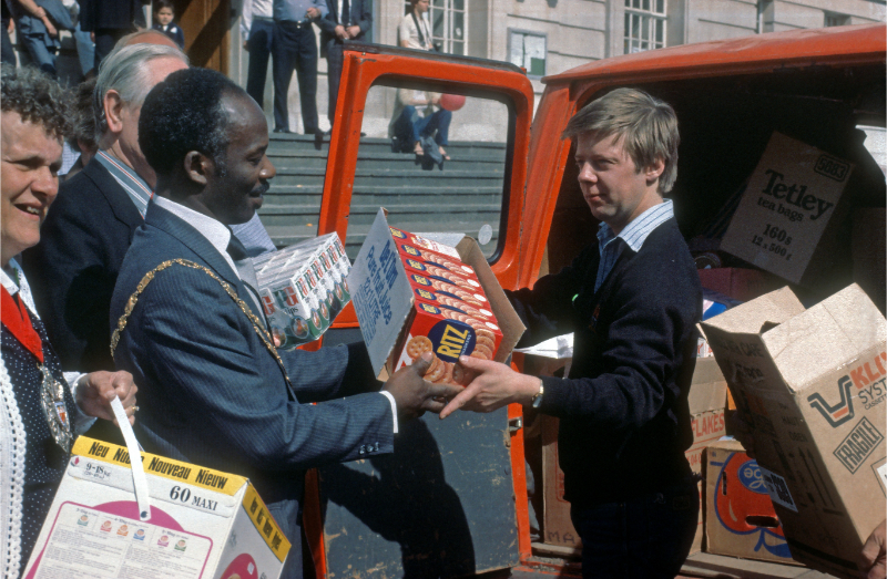 A colour photograph of a man wearing a suit and a gold ceremonial chain handing a box of crackers to a man next to an open red van parked in front of Hackney Town Hall.