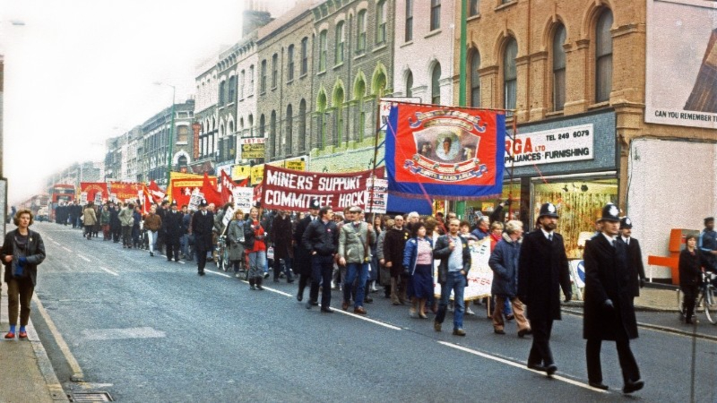 A colour photograph of a demonstration marching down Kingsland High Street, Hackney, in front of Centerprise Bookshop, with a police officer walking in front. Marchers hold banners for the National Union of Mineworkers South Wales Area, and the Miners Support Committee Hackney.