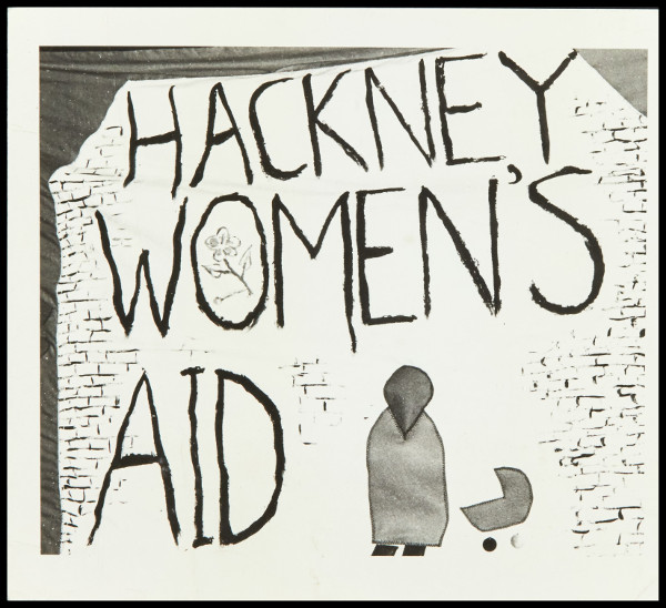 Black and white photo of a hand painted banner with the words 'Hackney Womens Aid' with a simple picture of a woman and a pram.