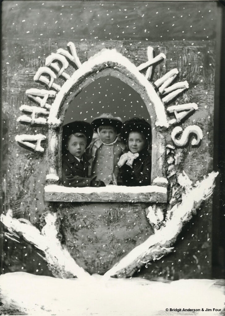 Black and white photograph three children posing in a Christmas card set - looking out of a window with 'A Happy Xmas' around the written around the window. Copyright to Bridgit Anderson and Jim Four.