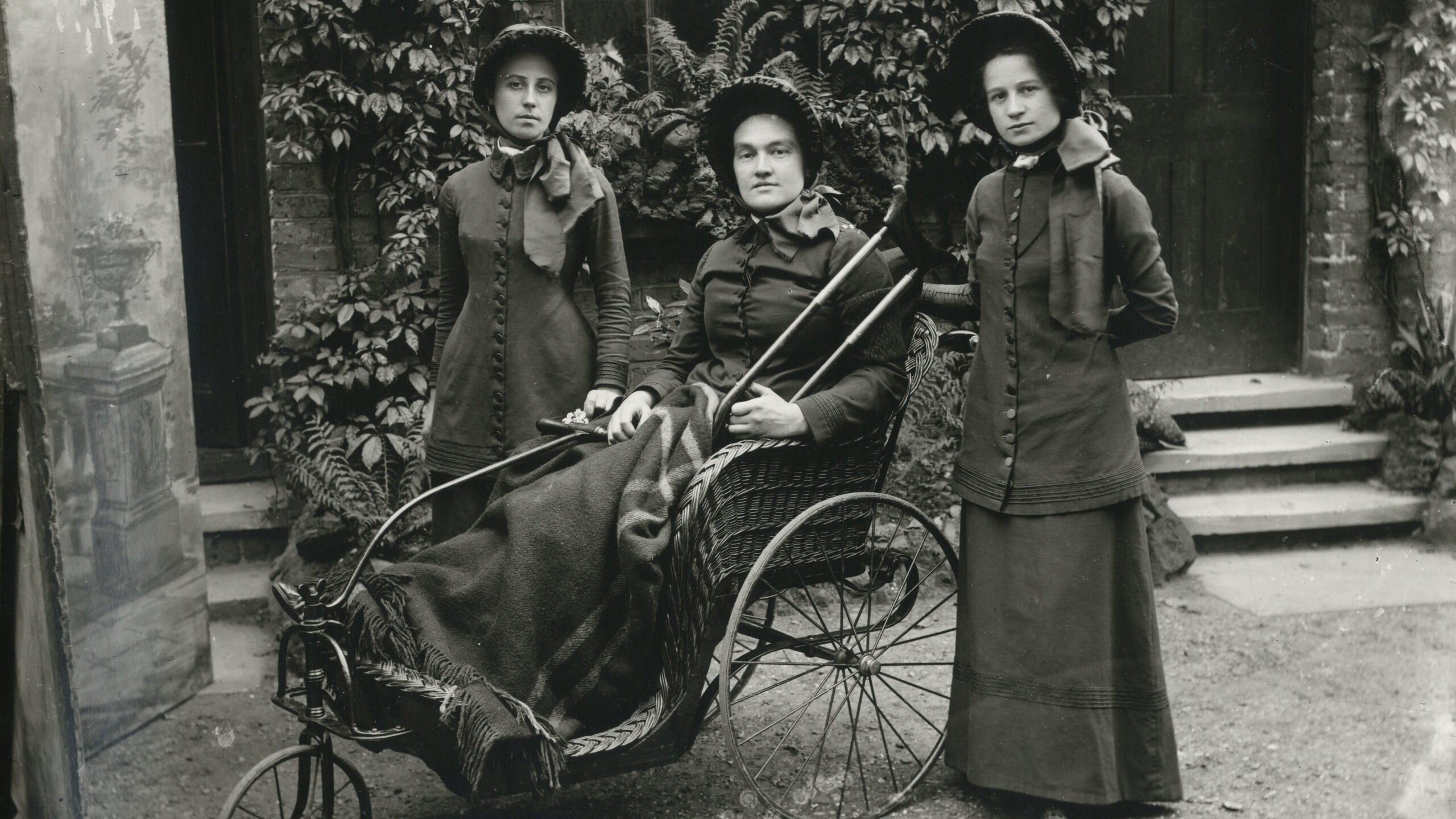 Black and white photograph of three women outside, dressed in traditional Victorian clothes and bonnets. One woman is a wheelchair user. Copyright to Bridgit Anderson and Jim Four.