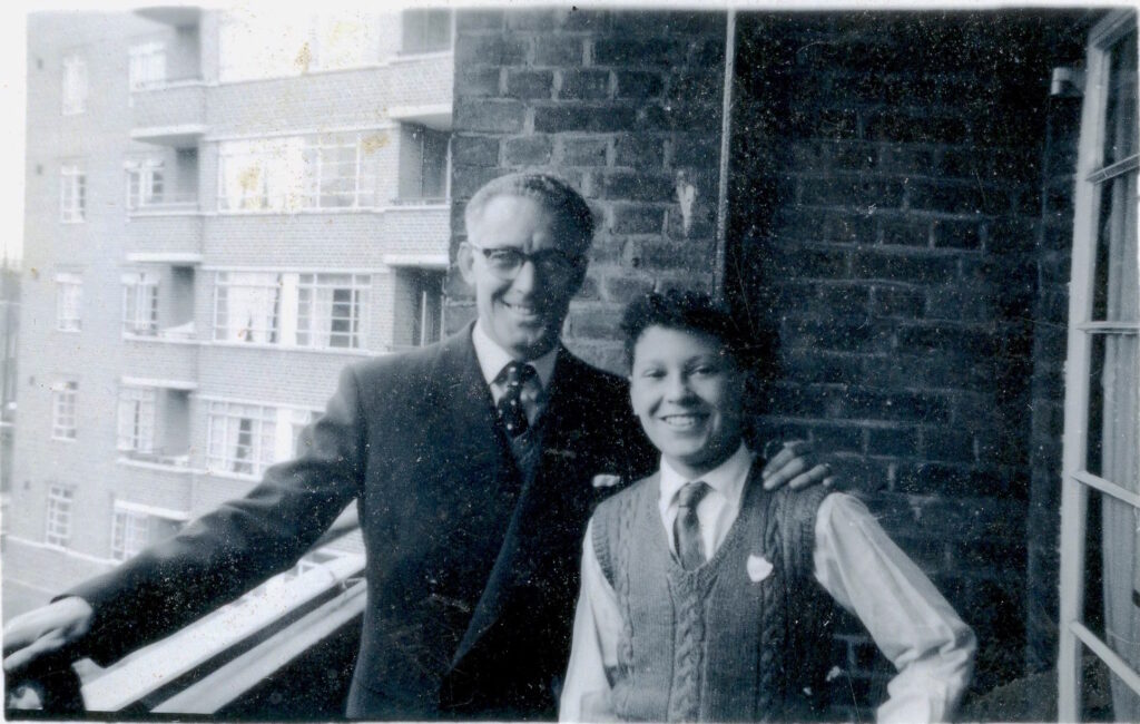 Black and white photo of two people standing on a walkway of a block of flats facing the camera and smiling, with a block of flats visible behind them. The man on the left is wearing glasses and a suit. The young boy on the right is wearing a tie, shirt and waistcoat.