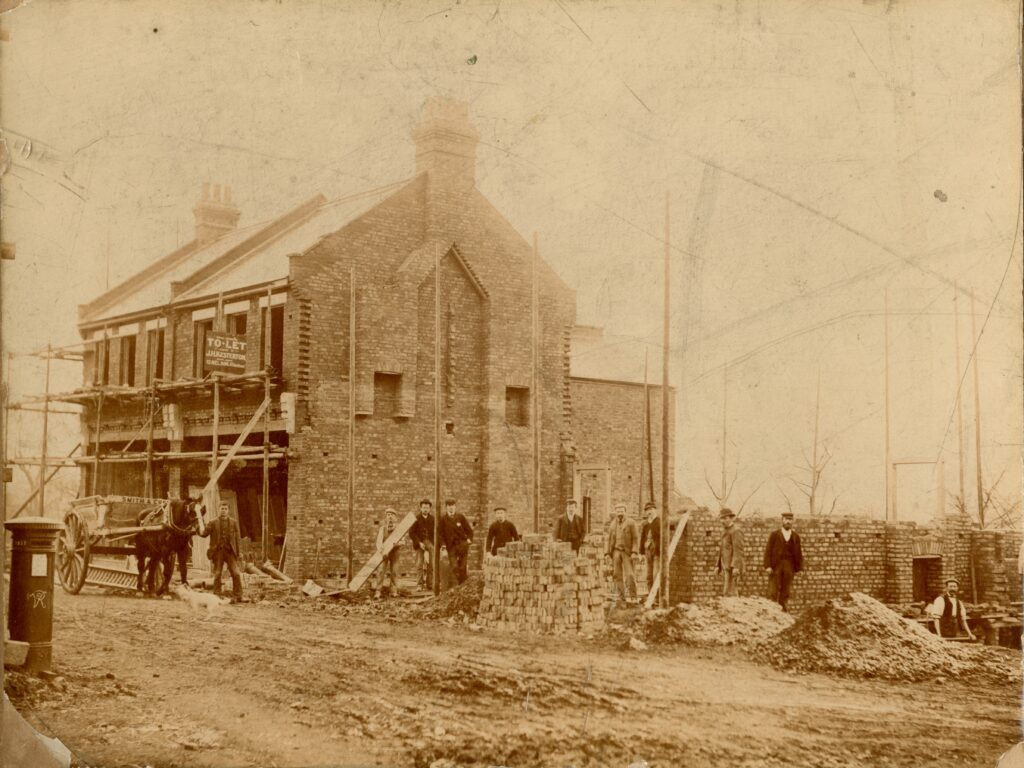 Historic, brown tinted photograph of a building site. The builders are posing and looking at the photographer. A horses and cart led by a man is also in the picture.