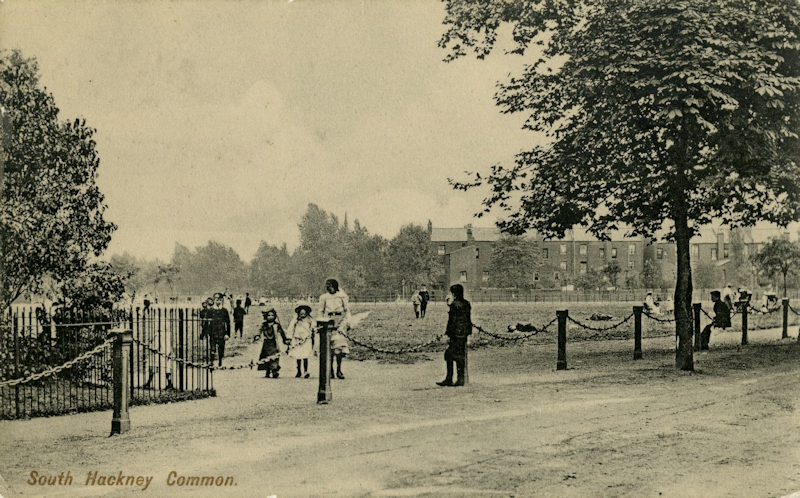 Sepia tinted view of an open space with grass and trees around the perimeter. People are enjoying the open space, including a woman with two young children in the foreground. There is a row of brick villas on the right of the image. The bottom left of the postcard says South Hackney Common.