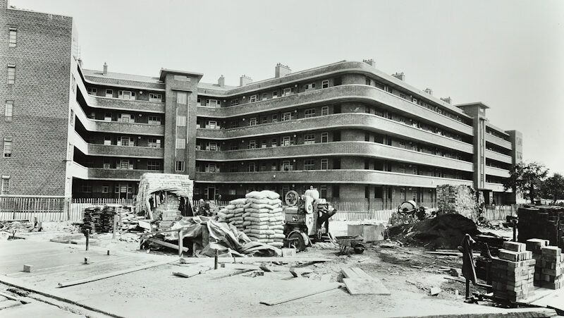 Black and white photograph showing a brick low rise housing estate with construction materials in front of it.