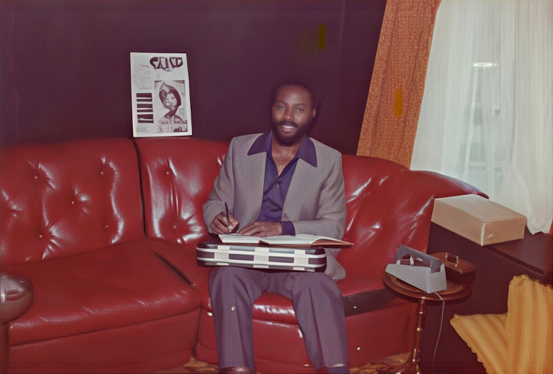A man seated on a red leather sofa with a closed briefcase resting on his knees and a notepad and diary open on top. He is holding a pen to write on these, but looking at the camera to be photographed.