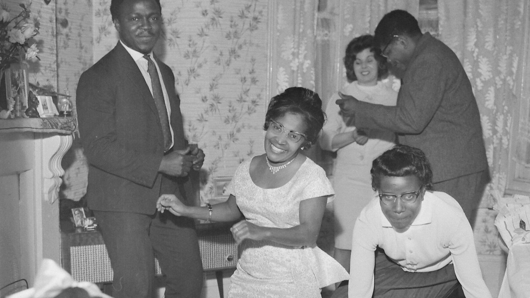 Black and white photograph of a Caribbean family dancing around a radiogram in their home.