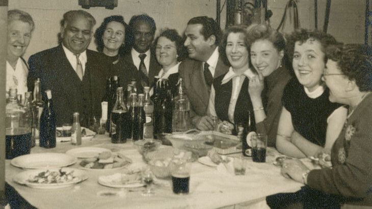 Black and white photograph showing a group of 10 adults, 3 men and 7 women, seated around a table looking towards the camera. The table has an assortment of drinks and food.