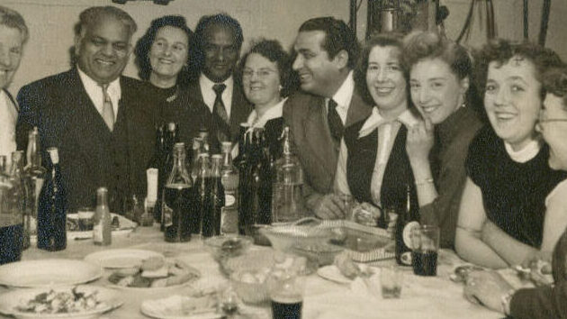Black and white photograph showing a group of 10 adults, 3 men and 7 women, seated around a table looking towards the camera. The table has an assortment of drinks and food.