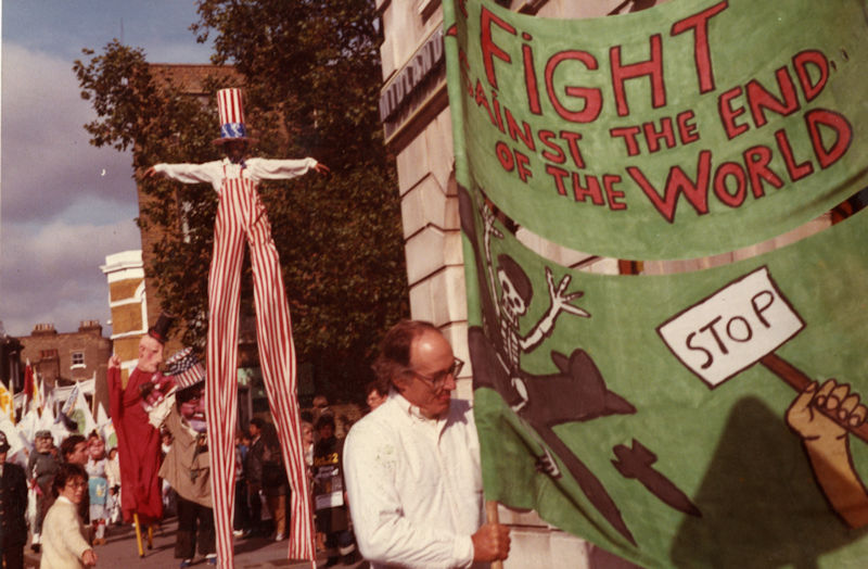 A photograph of a Peace Carnival in Hackney in 1983, In the foreground, a large green banner held by a man reads “FIGHT AGAINST THE END OF THE WORLD” with an illustration of a skeleton. In the background, in front of a crowd, a performer wearing red and white striped clothing and a matching large hat, stands on stilts.