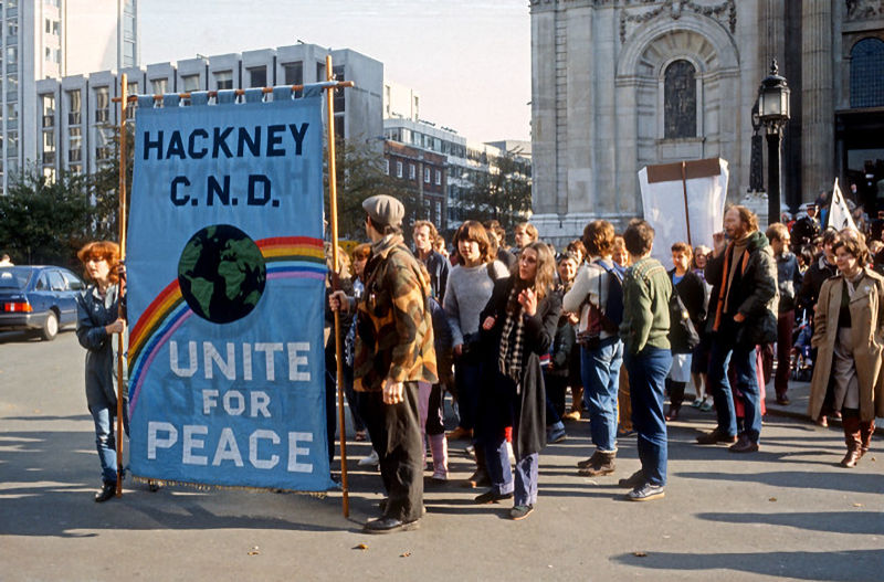 Protesters lined up in an urban area holding a sign with the earth and a rainbow reading “Hackney C.N.D. Unite for peace.”