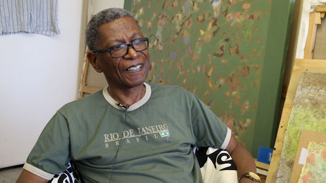 Man sitting in his artists studio, wearing a green t-shirt, with large canvases of his paintings behind him.