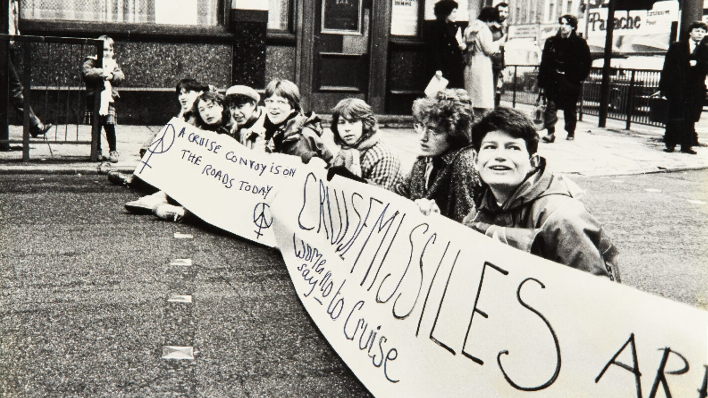 A photograph of a group of women protesting on the street while holding a large banner with large text about cruise missiles. The banner is held by the group and is wide enough to cover the road they are sitting on.